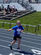 A young female student running down a blue all-weather track during a race. She is wearing a blue t-shirt and a competition bib, with spectators and hurdles visible in the background.